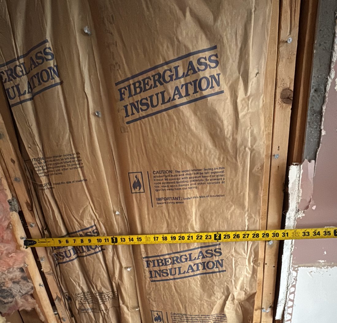 Kraft-faced fiberglass batt insulation installed in an open wall with a tape measure across the studs